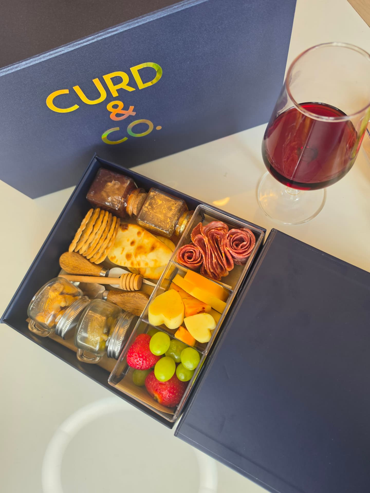 Curd & Co. navy signature box with cheeses, salami roses, fruit and a glass of red wine