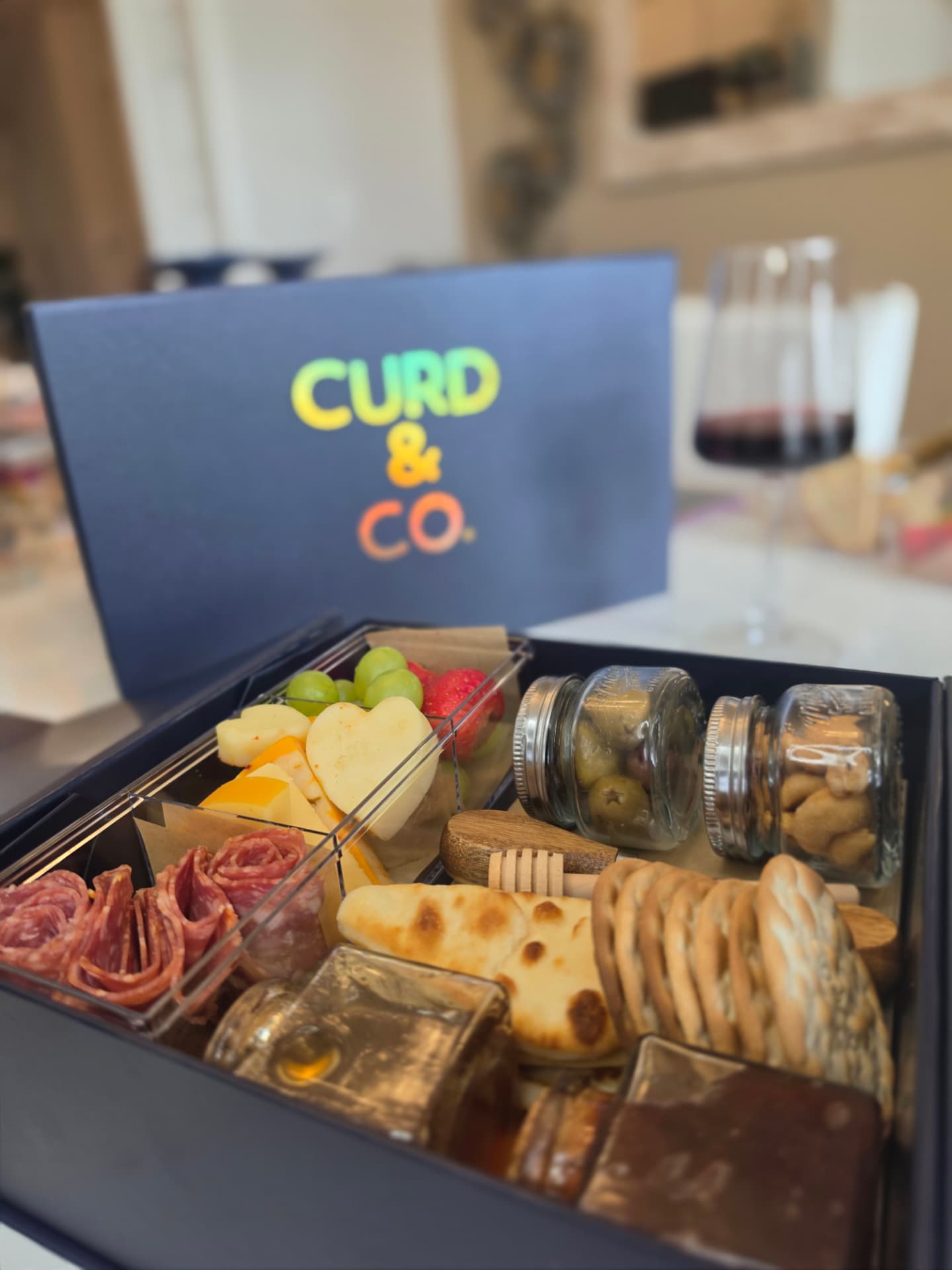Navy Curd & Co. box open with heart cheeses, salami, olives and honey beside a glass of red wine