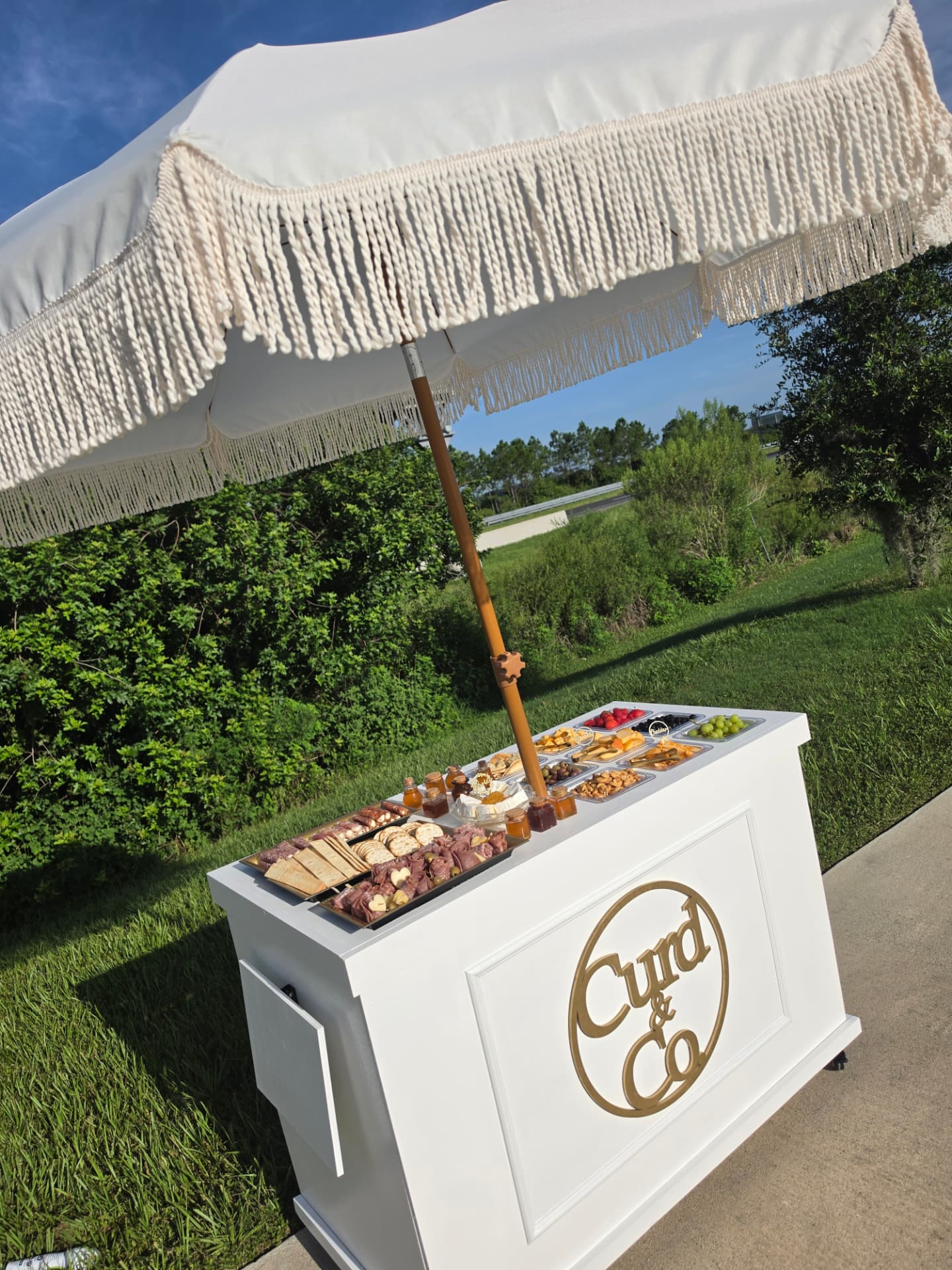 Curd & Co. branded mobile charcuterie bar under a fringed umbrella